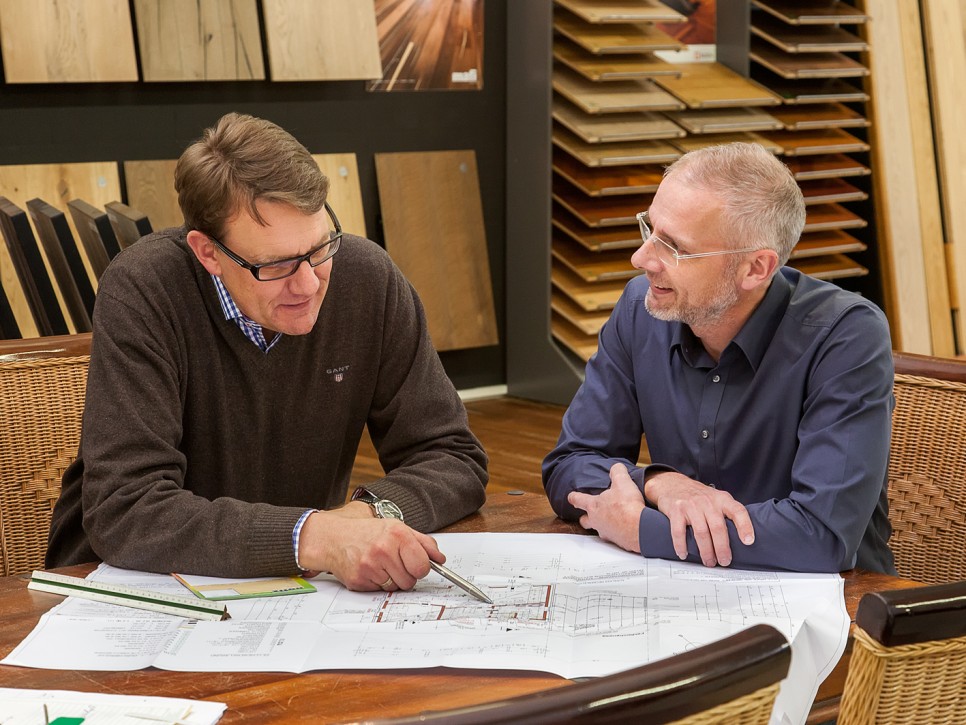 Beratungsgespräch über Bodenbelagsplanung in einer Ausstellung Zwei Personen bei der Besprechung von Bauplänen in einer Bodenbelag-Ausstellung mit Parkettmustern im Hintergrund
