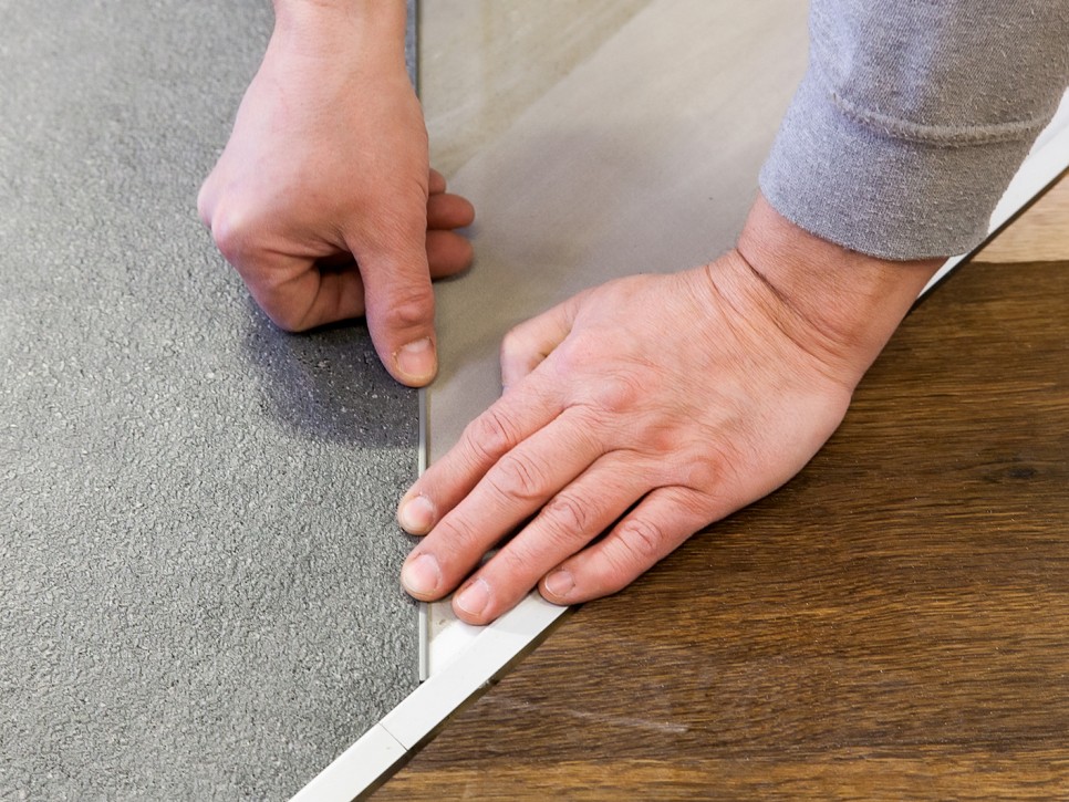 Einpassen einer Bodenleiste zwischen verschiedenen Bodenbelägen Nahaufnahme von Händen beim Montieren einer Übergangsleiste zwischen grauem Belag und Holzoptikboden