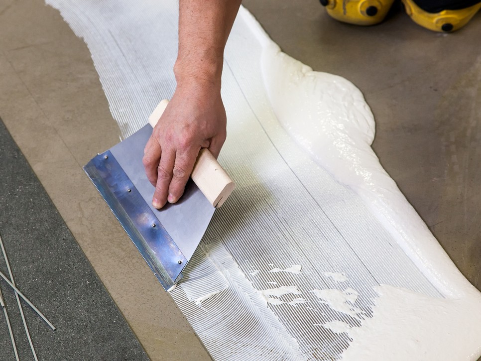 Auftragen von Kleber mit einer Zahnkelle bei der Bodenverlegung Nahaufnahme einer Hand, die mit einer Zahnkelle weißen Kleber gleichmäßig auf dem Untergrund verteilt